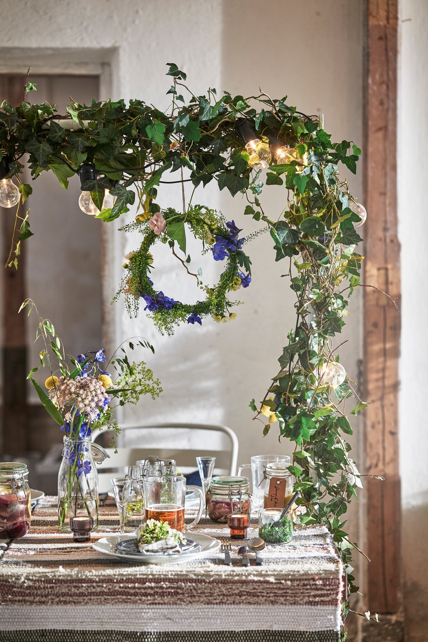 En festlig dukning med ett rustikt bord under en vacker båge av murgröna och en hängande krans av blåa blommor.