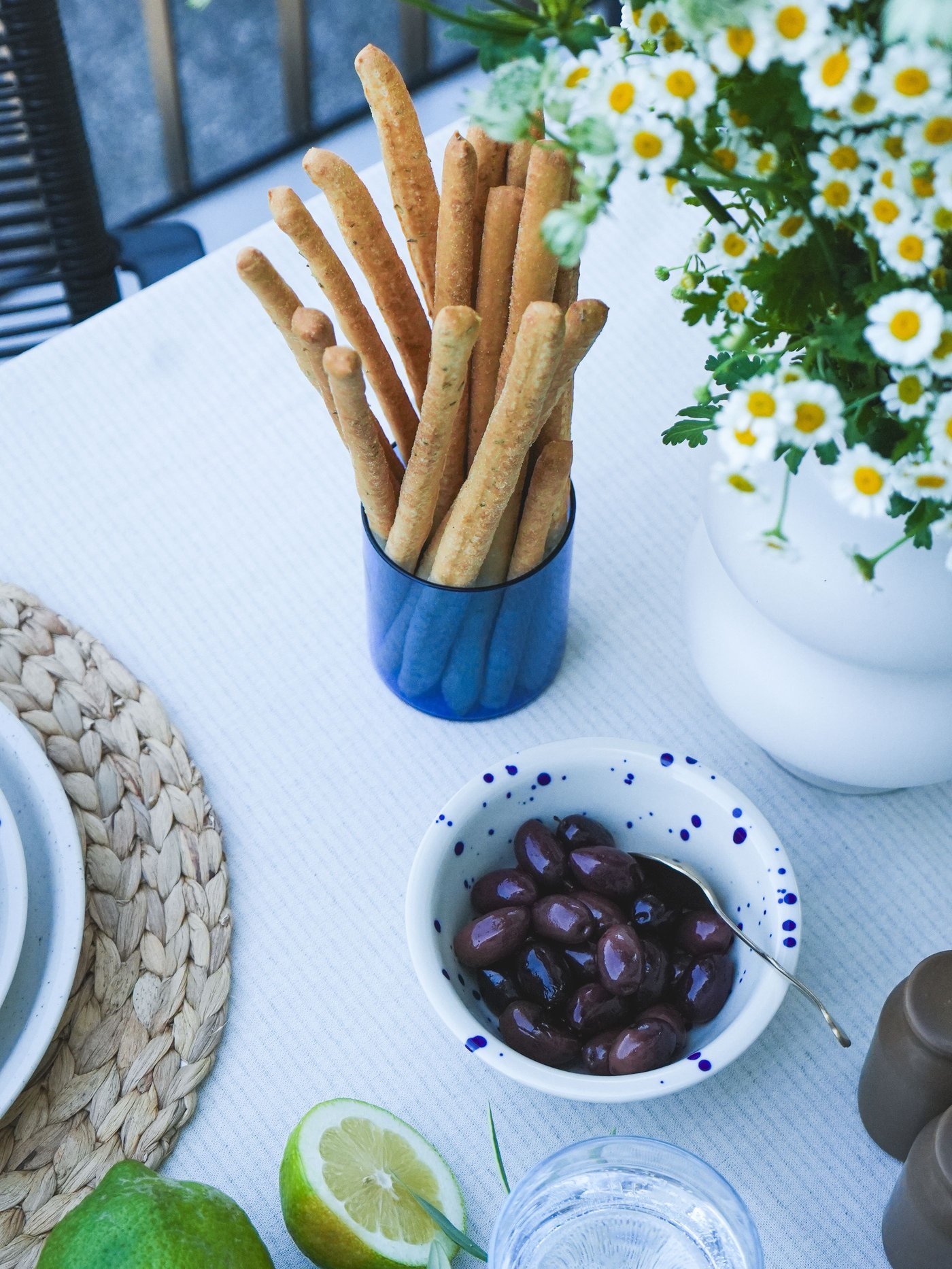 Gedeckter Tisch mit Grissini in einem blauen Glas, einer SKUGGSTUBB Becher mit weissen Gänseblümchen, einer SILVERSIDA Schale mit schwarzen Oliven, frischen Zitronen und Limetten.