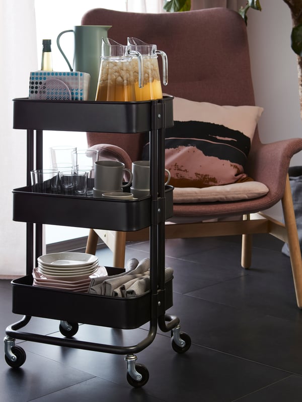 A white RÅSKOG trolley containing 4 storage boxes with baby towels, hairbrush, bottle and nappies on a brown carpet flooring.