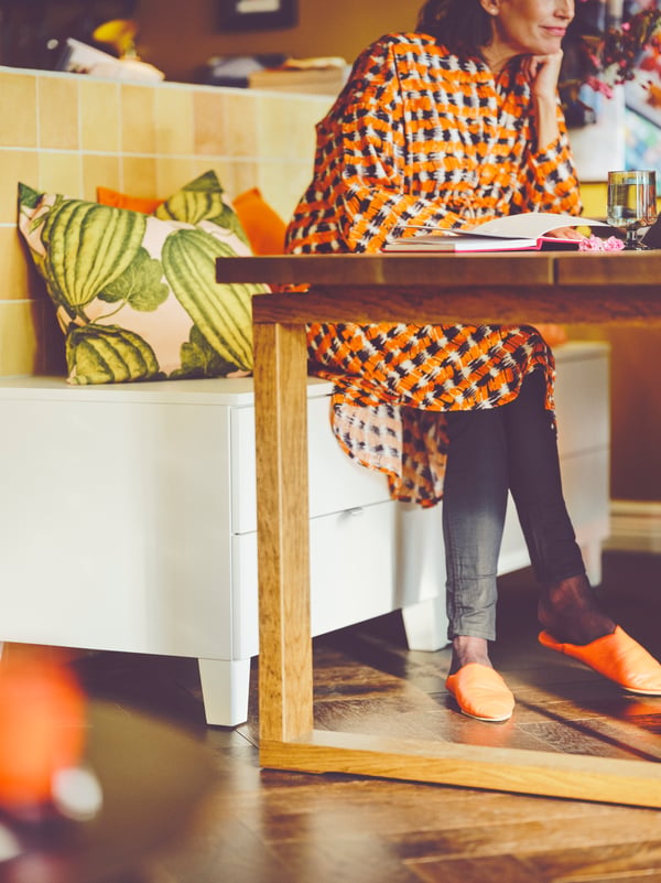 An adult sits on a white storage bench at a wooden MÖRBYLÅNGA dining table. Cushions sit behind her.