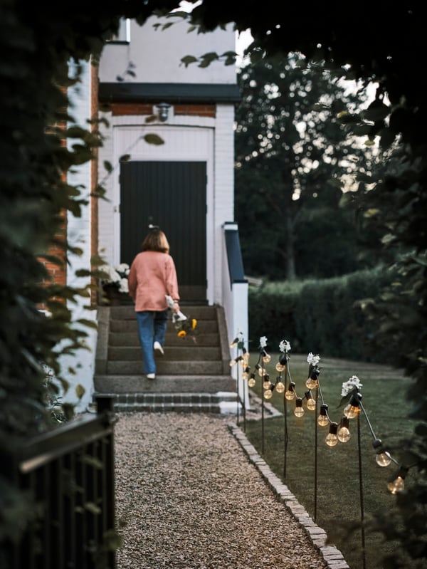 Black SVARTRÅ LED lighting chains hanging along a gravel path leading up to a house where a person is walking up the stairs.