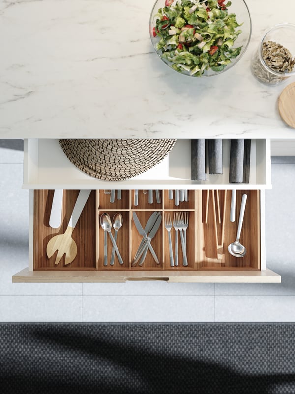 Vista superior de una cubierta laminada EKBACKEN efecto mármol blanco. Se ve un cajón del mueble inferior abierto con organizadores de cubiertos de color madera clara.