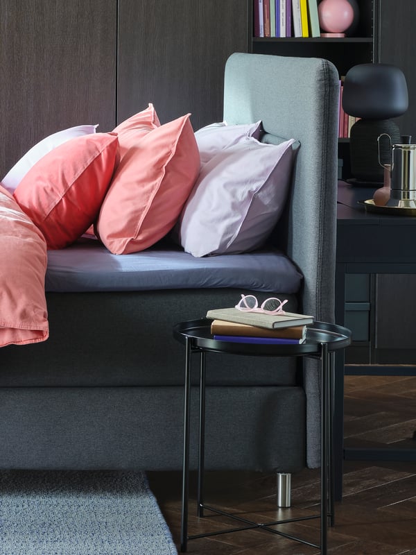 A grey FINNSNES divan bed with bed linen including light brown-red ÄNGSLILJA pillowcases next to a GLADOM tray table.