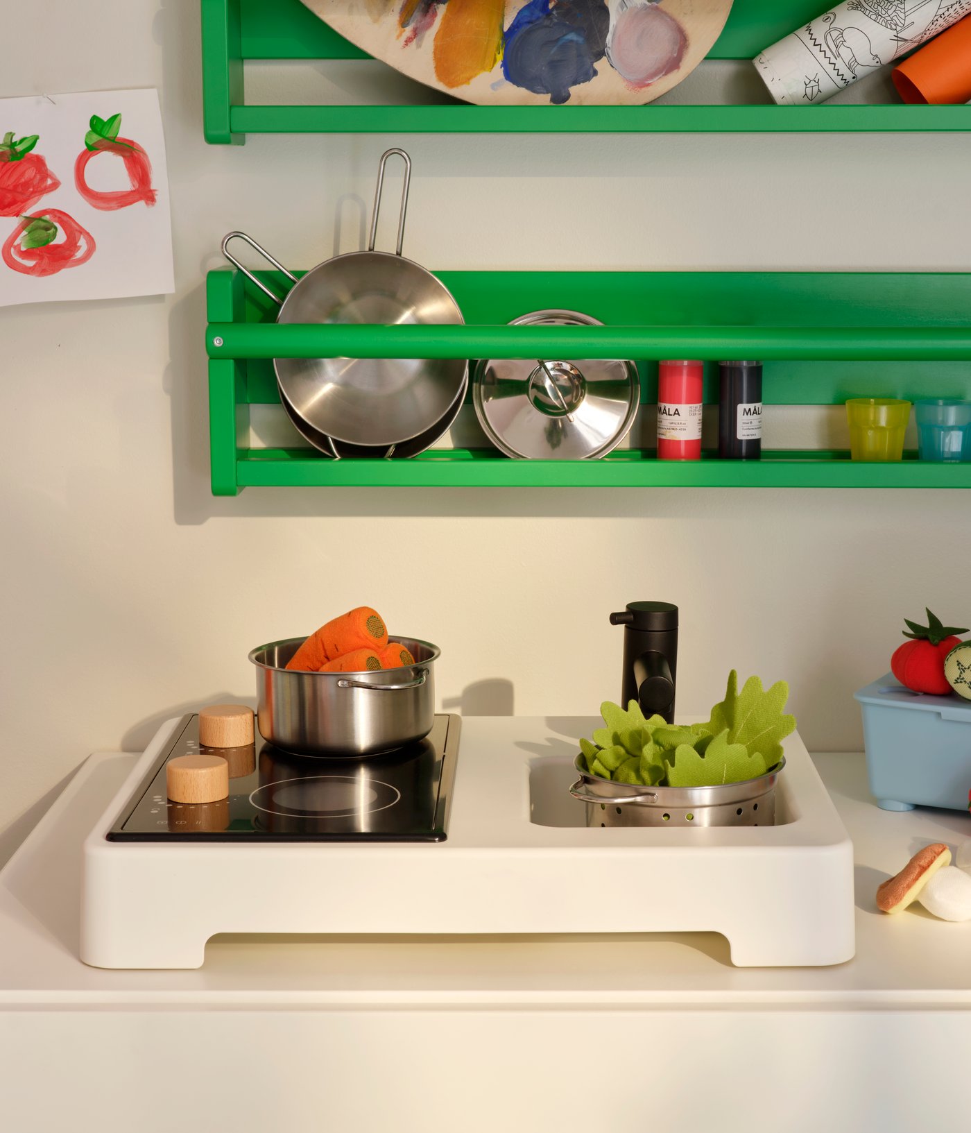 Parts of a STENMUS mini-play kitchen beneath green FLISAT wall storage, all filled with play-cooking accessories.