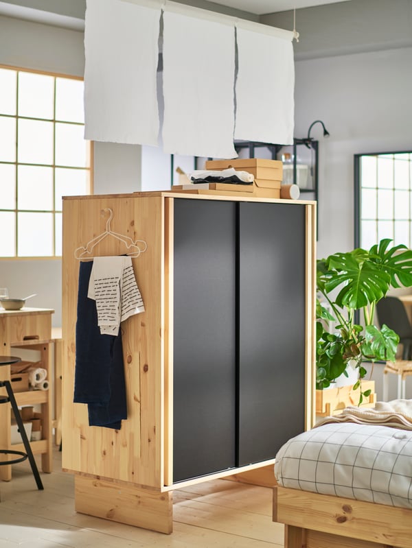 GLAMBERGET wardrobe in untreated wood with black sliding doors, placed in the middle of the room as a room divider.