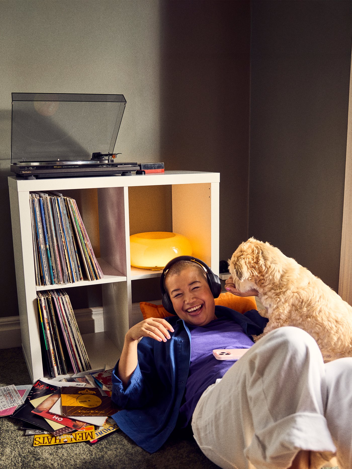 A cozy scene features a person relaxing with headphones while a dog curiously leans in, surrounded by a record player and a shelf filled with vinyl records.