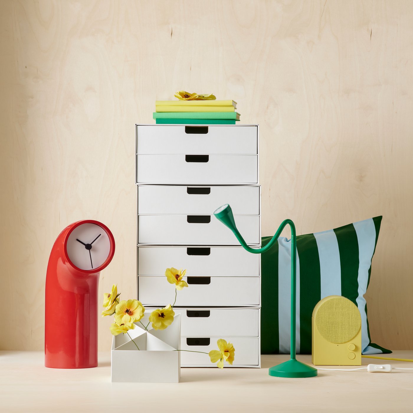 Productos en un estudio, incluidos una cómoda blanca, un reloj de mesa rojo, una lámpara de escritorio verde y flores artificiales amarillas.