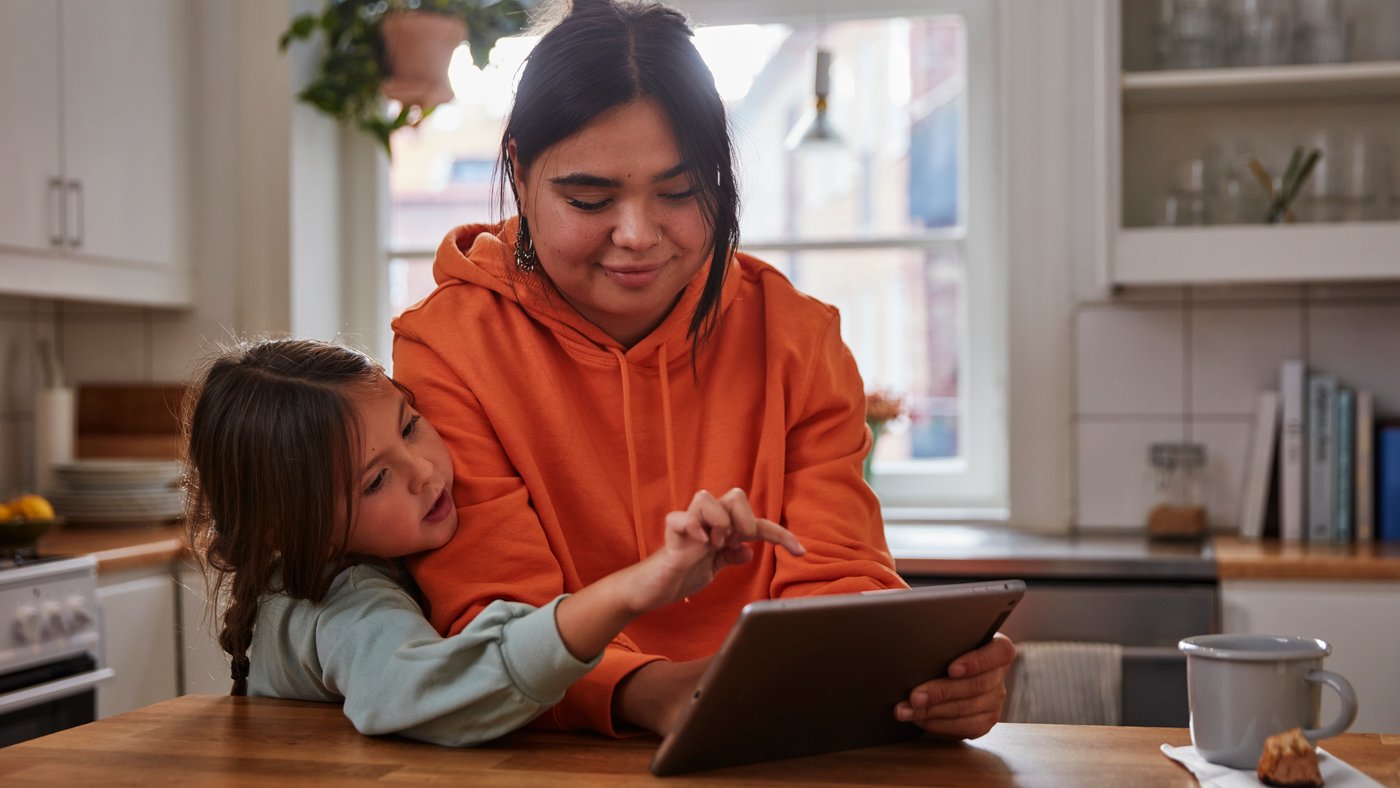 A child and an adult sitting together at a kitchen table, using a tablet, with morning light coming through the window and a warm, homey atmosphere around them.