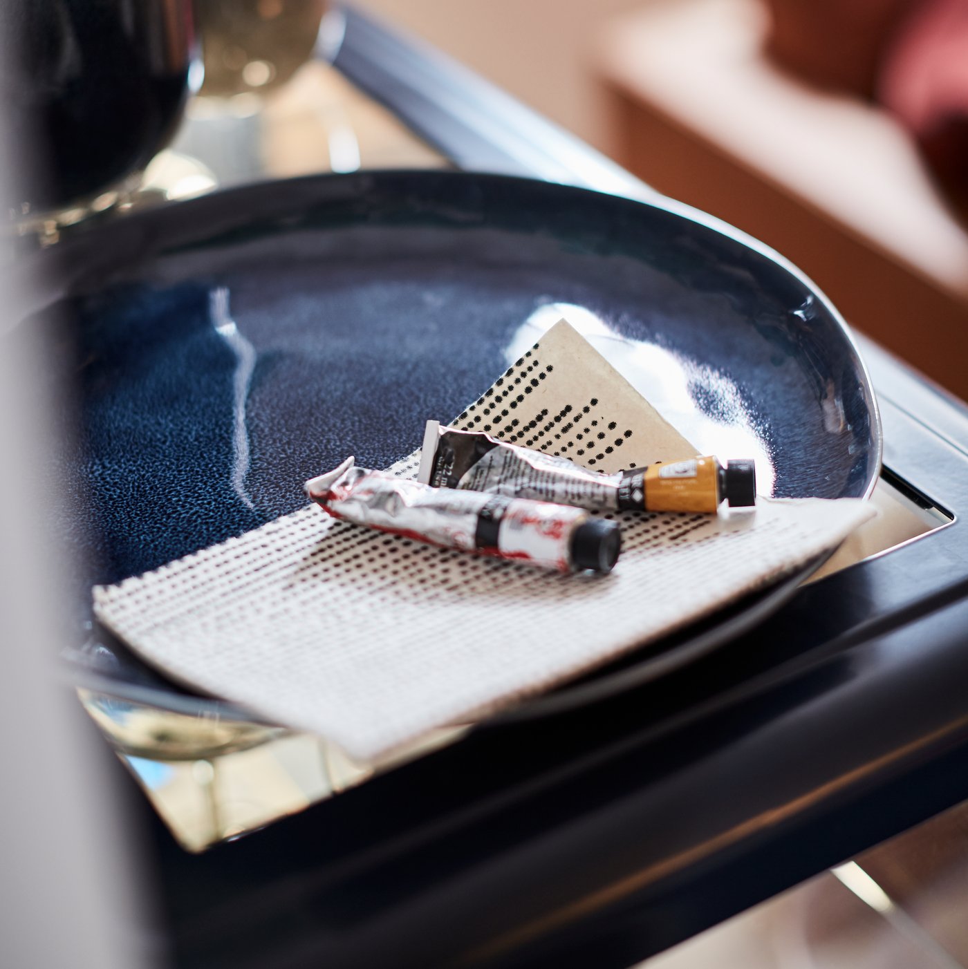 A close-up of two watercolour paint tubes and a piece of paper lying in an ERTAPPAD dish in blue on a table corner.