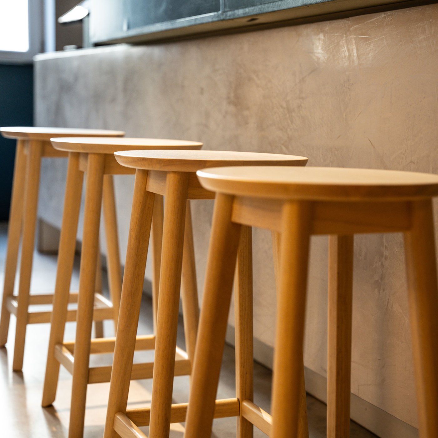 Rij houten barkrukken aan lichte toonbank met betonlook. Minimalistisch design in modern interieur met warme lichtinval en donkere achterwand.