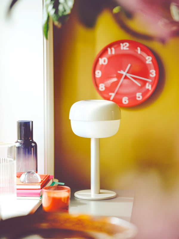 A beige BLÅSVERK table lamp and red PLUTTIS wall clock are set off by a mustard wall. Coloured glass items sit nearby.