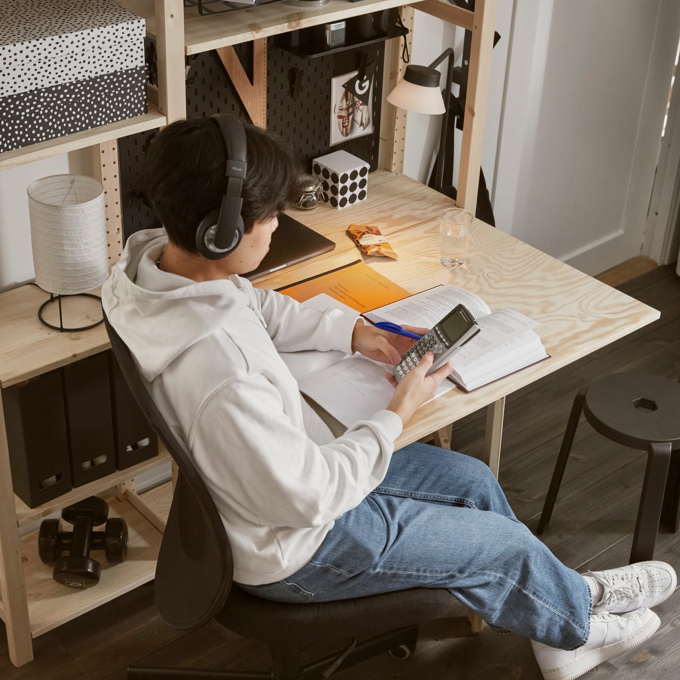 A person is studying with books on a black office chair at an IVAR desk, which is used as a drop‑leaf table attached to an IVAR shelving system, all made of pine.