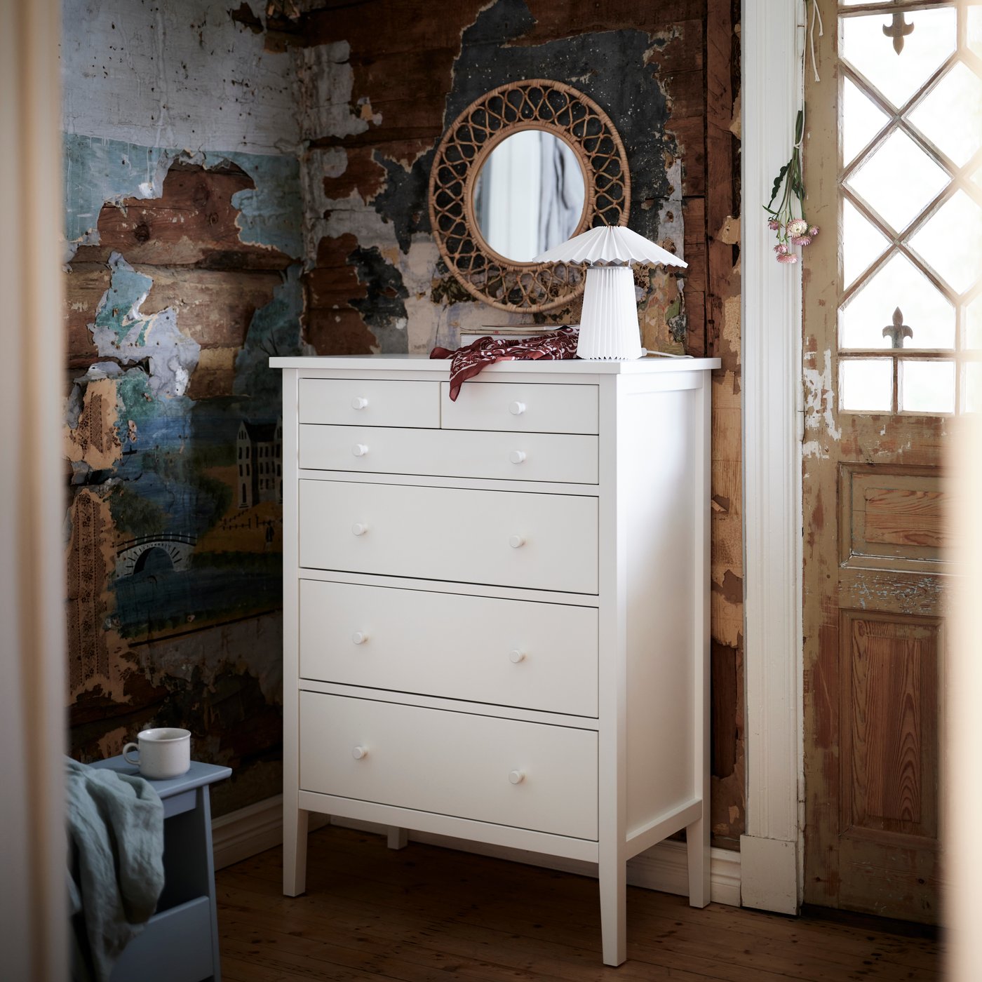 A white GULLABERG chest of 6 drawers stands against a rustic wall with a round mirror, lamp and small items arranged on top.