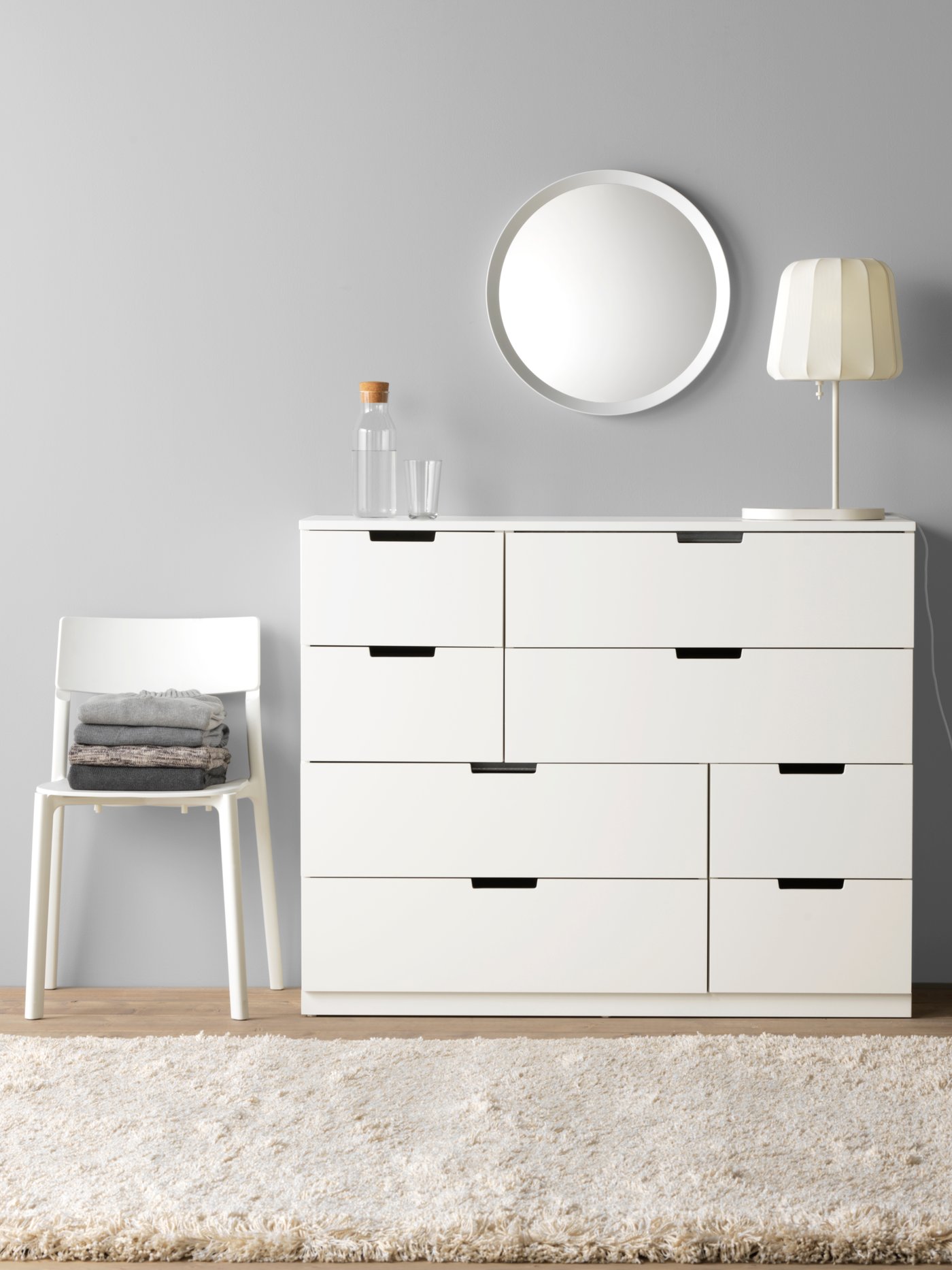 A white chest of drawers with black handles stands against a grey wall, with a round mirror and lamp on top, and a white chair with folded clothes beside it.