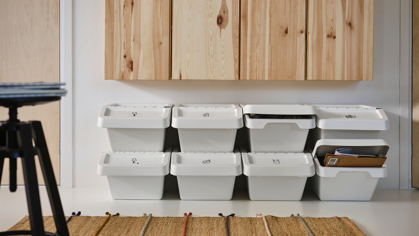 Rows of white SORTERA waste sorting bins with lids stacked on top of one another under a row of pine cabinets, plus a rug.