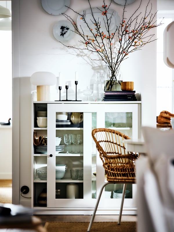 A white HAUGA cabinet with bi-fold glass doors in a white country-style room filled with objects, by a NILSOVE armchair.