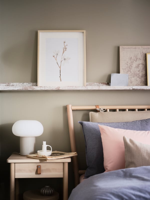 A BJÖRKSNÄS birch bedside table with a white DEJSA lamp on it stands beneath a picture ledge with art, and beside a bed.