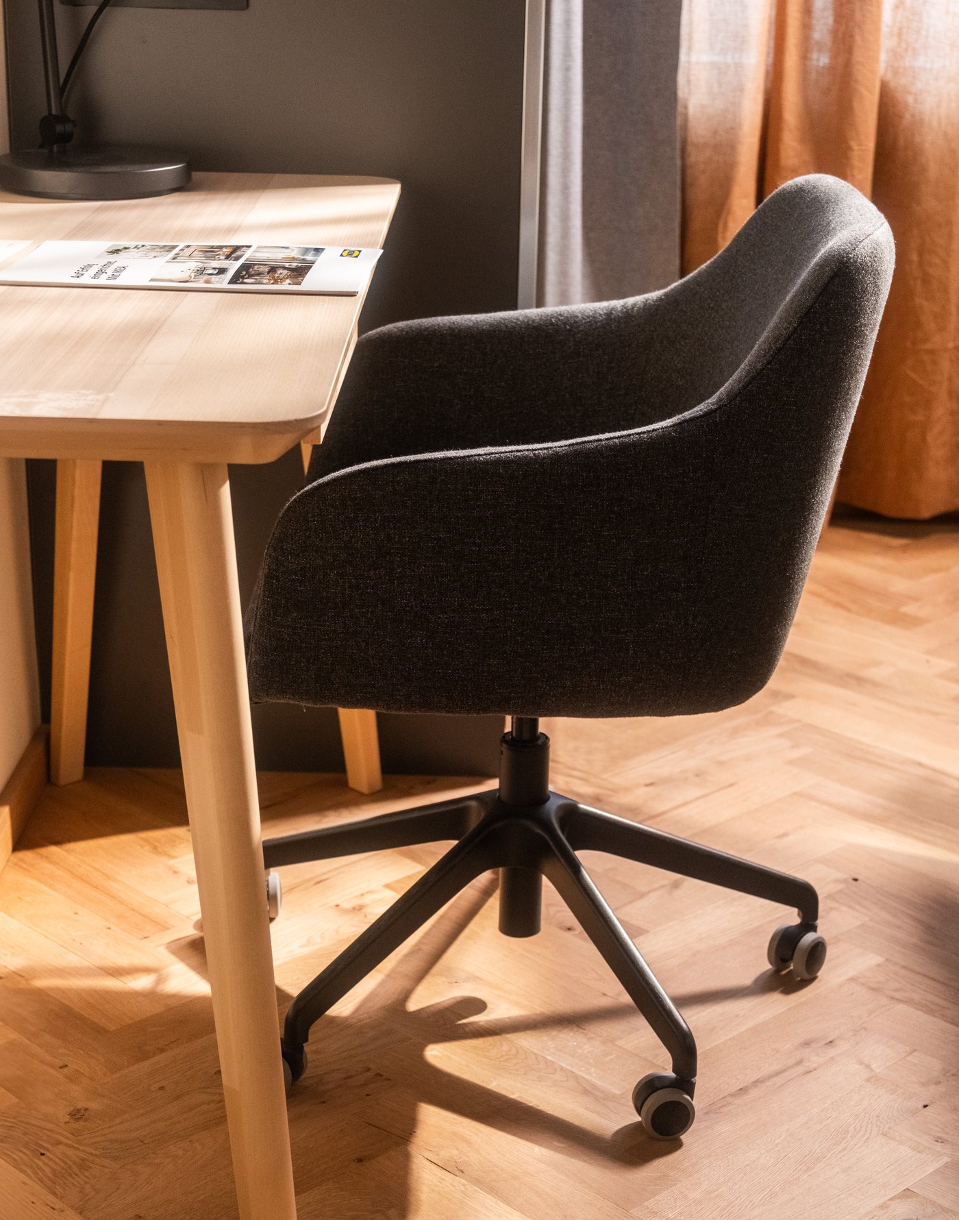 Dark upholstered desk chair on wheels at a light wooden table with an open newspaper, in warm lighting and wooden floors.