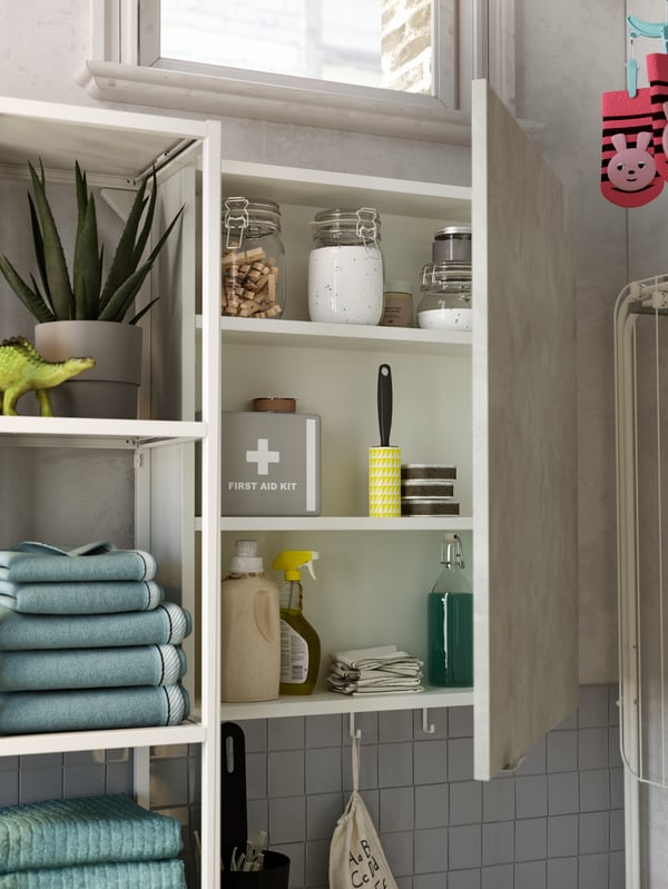 A narrow cabinet with its door open showing a first aid kit, laundry detergent in jars, pegs and other cleaning items.