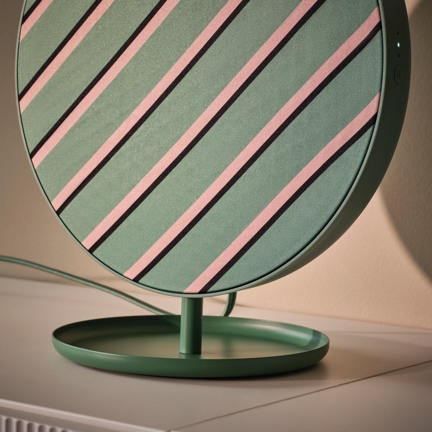 A green SOLSKYDD Bluetooth speaker with a striped design and mounted on its stand, placed on a surface by a wall.