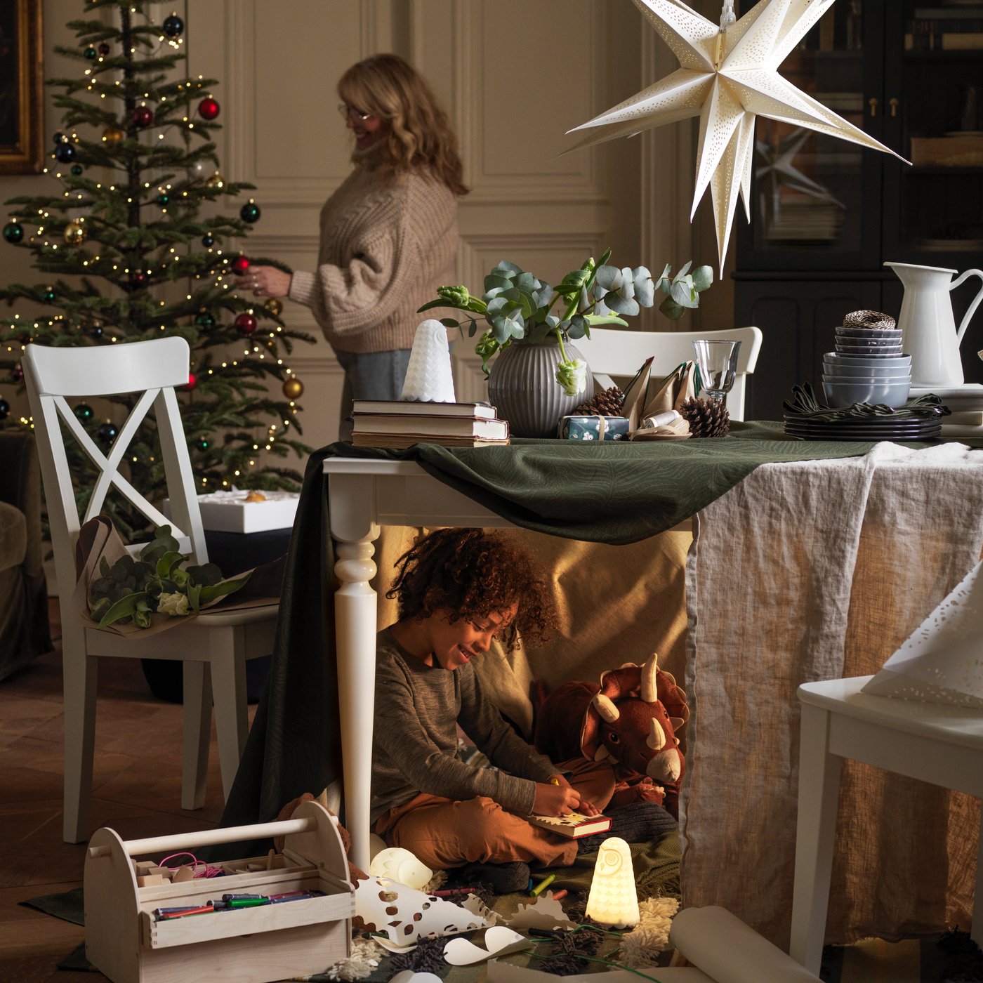 Une personne décore le sapin de Noël et sous la table au premier plan, un enfant joue à moitié caché avec des jouets.