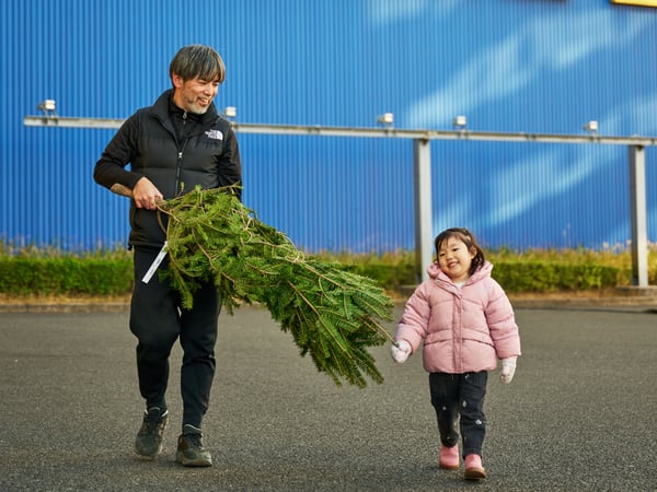 駐車場で、男性と女の子が笑顔でクリスマスツリーを運んでいます。背景にはIKEAの建物が見えます。