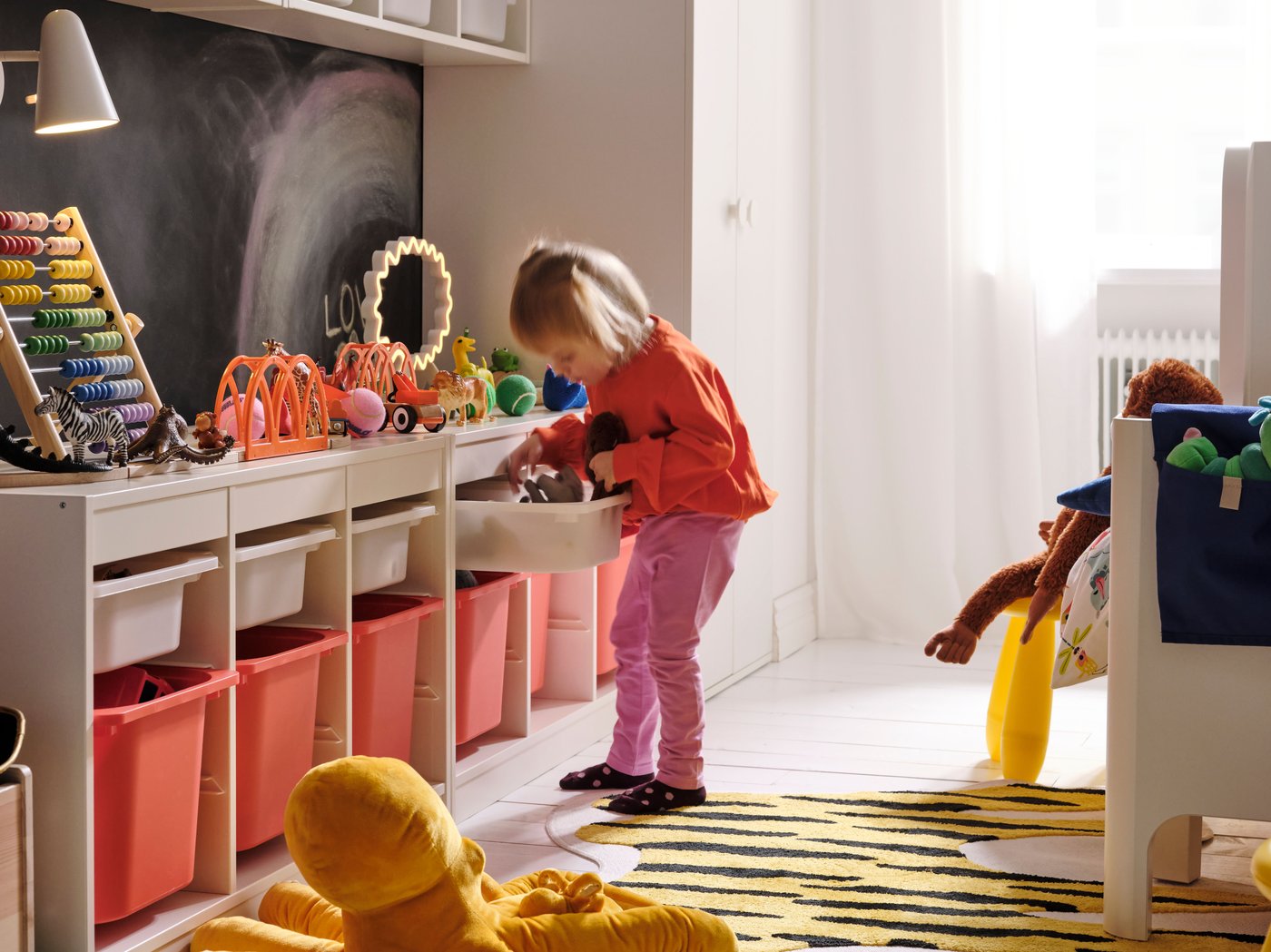 Dans une chambre d’enfant, une fillette farfouille dans l’un des bacs d’une longue combinaison de rangements pour jouets installée le long du mur.