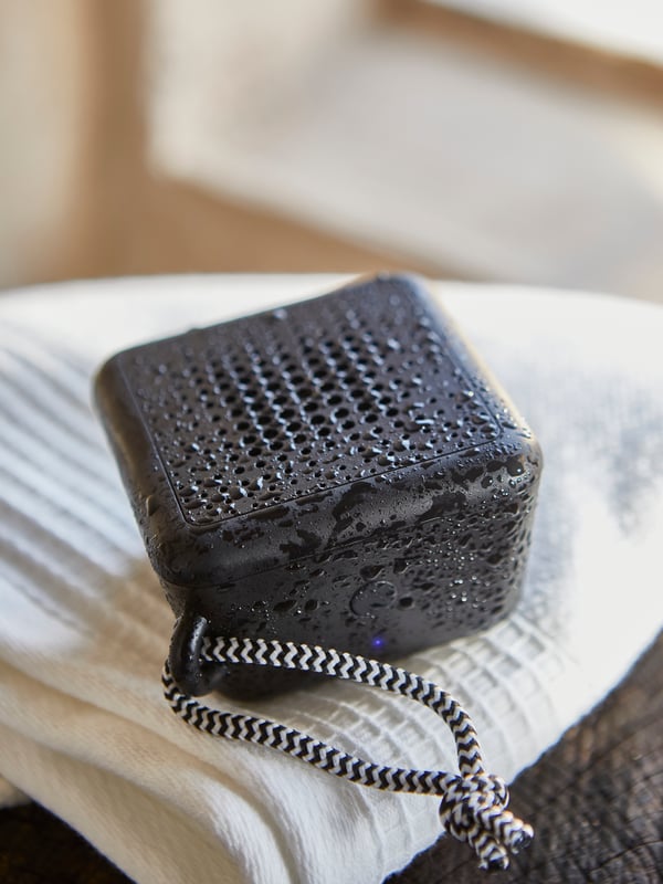 A waterproof black VAPPEBY portable Bluetooth speaker, covered in water drops, has been placed on a white VINARN bath towel.