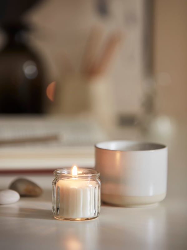 A lit Scandinavian Woods/white ADLAD scented candle in glass and a stoneware mug on a white KALLAX desk.
