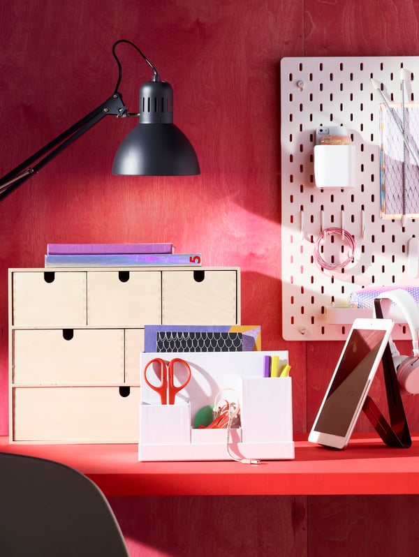 Su uno sfondo rosso, una scrivania rossa su cui ci sono diversi prodotti IKEA, tra cui una lampada, un pannello portaoggetti, una minicassettiera, forbici e penne.