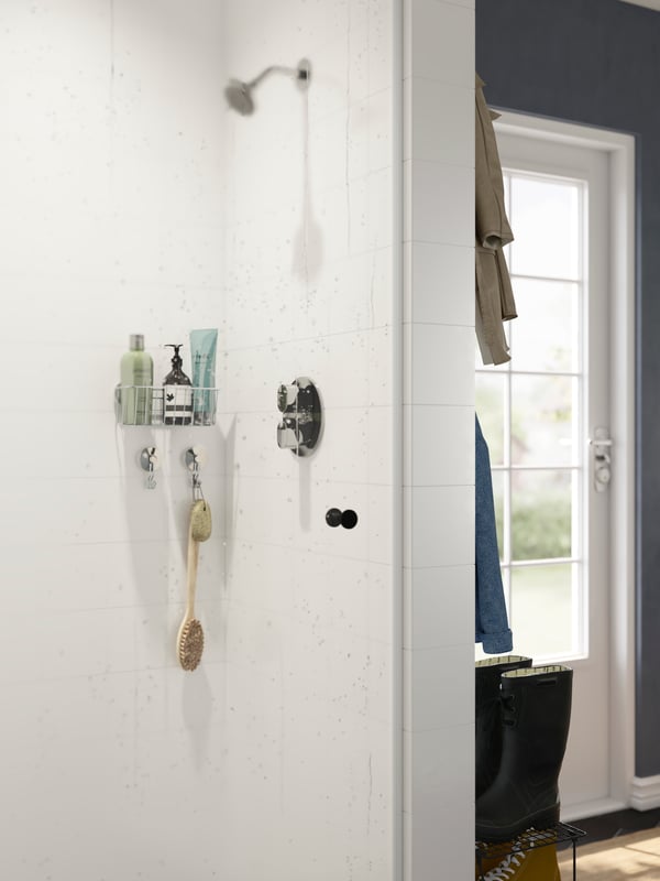 A shower is attached to a wall in a corner of a mudroom. There is a shelf with bathroom products and a brush on the wall.