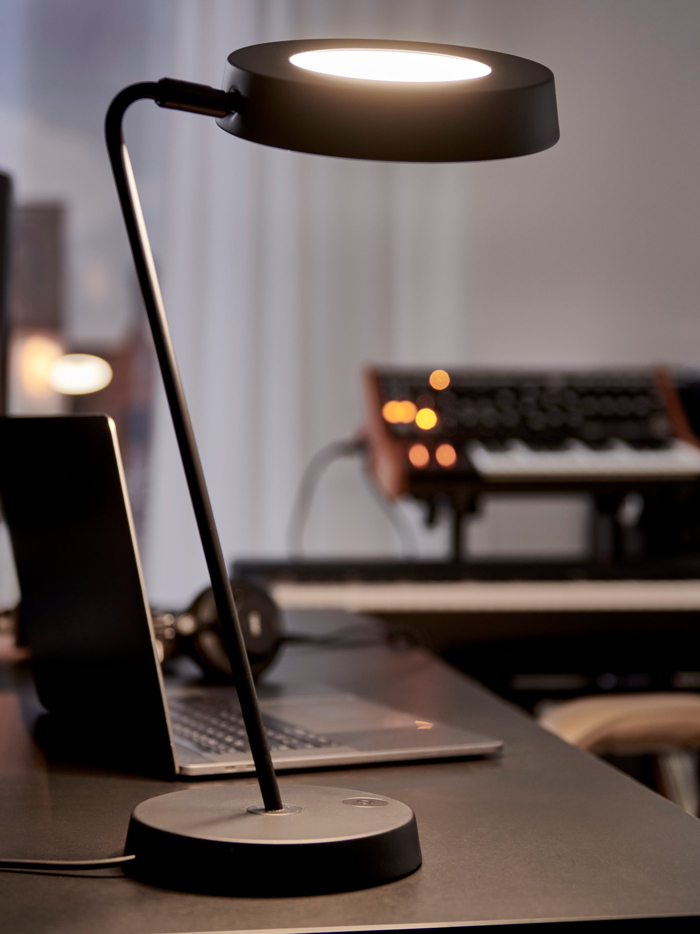 Rozsvícená černá pracovní lampa LED OBEGRÄNSAD na černém stole vedle otevřeného notebooku s klávesami v pozadí.