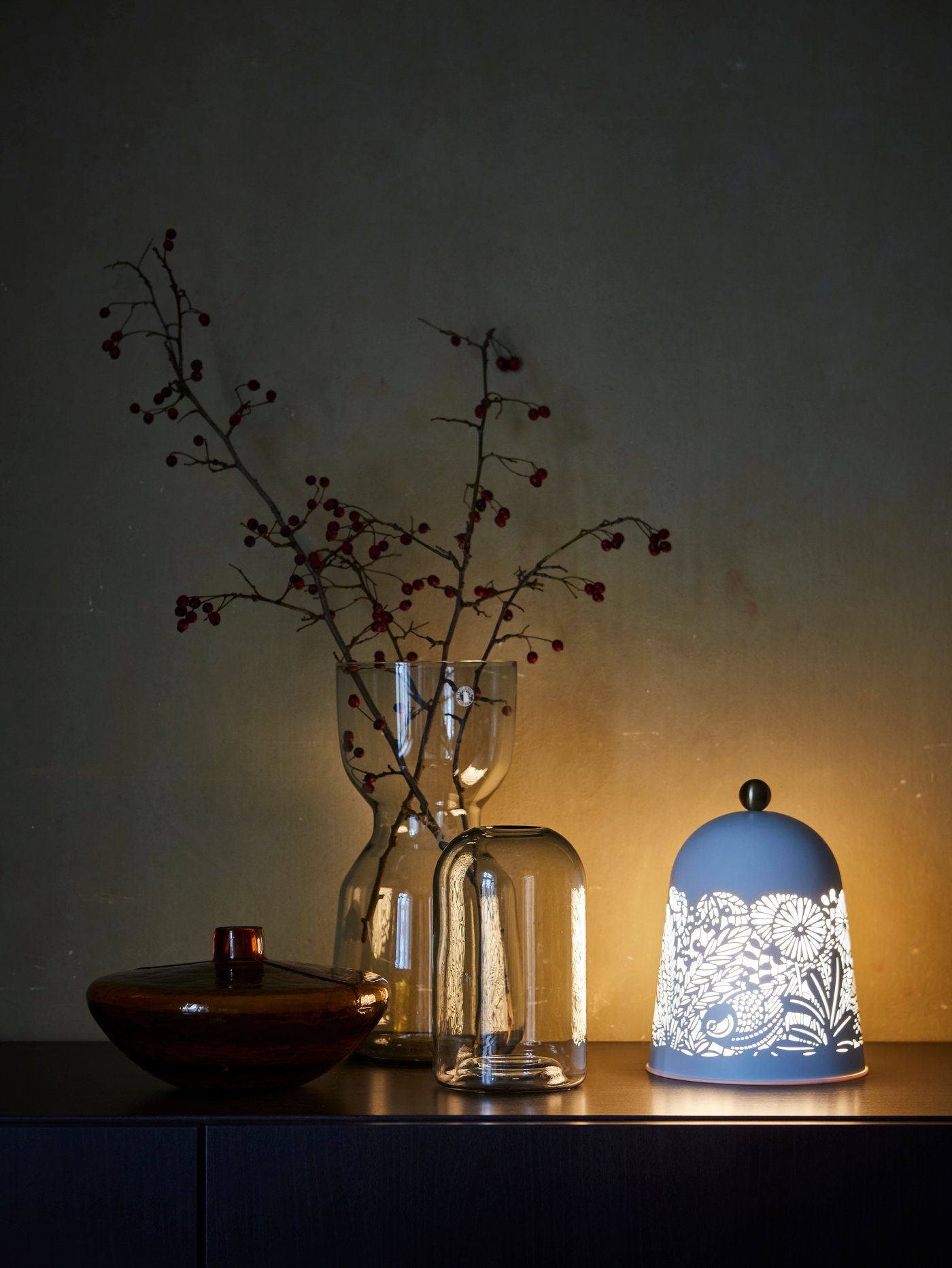 A tall decorative vase with autumn foliage, two short vases and a nature-inspired table lamp set on a sideboard.