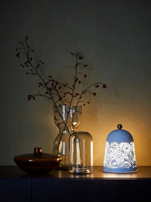 Eine hohe dekorative Vase mit Herbstlaub, zwei kleine Vasen und einer Tischleuchte mit Naturmotiv stehen auf einem Sideboard.