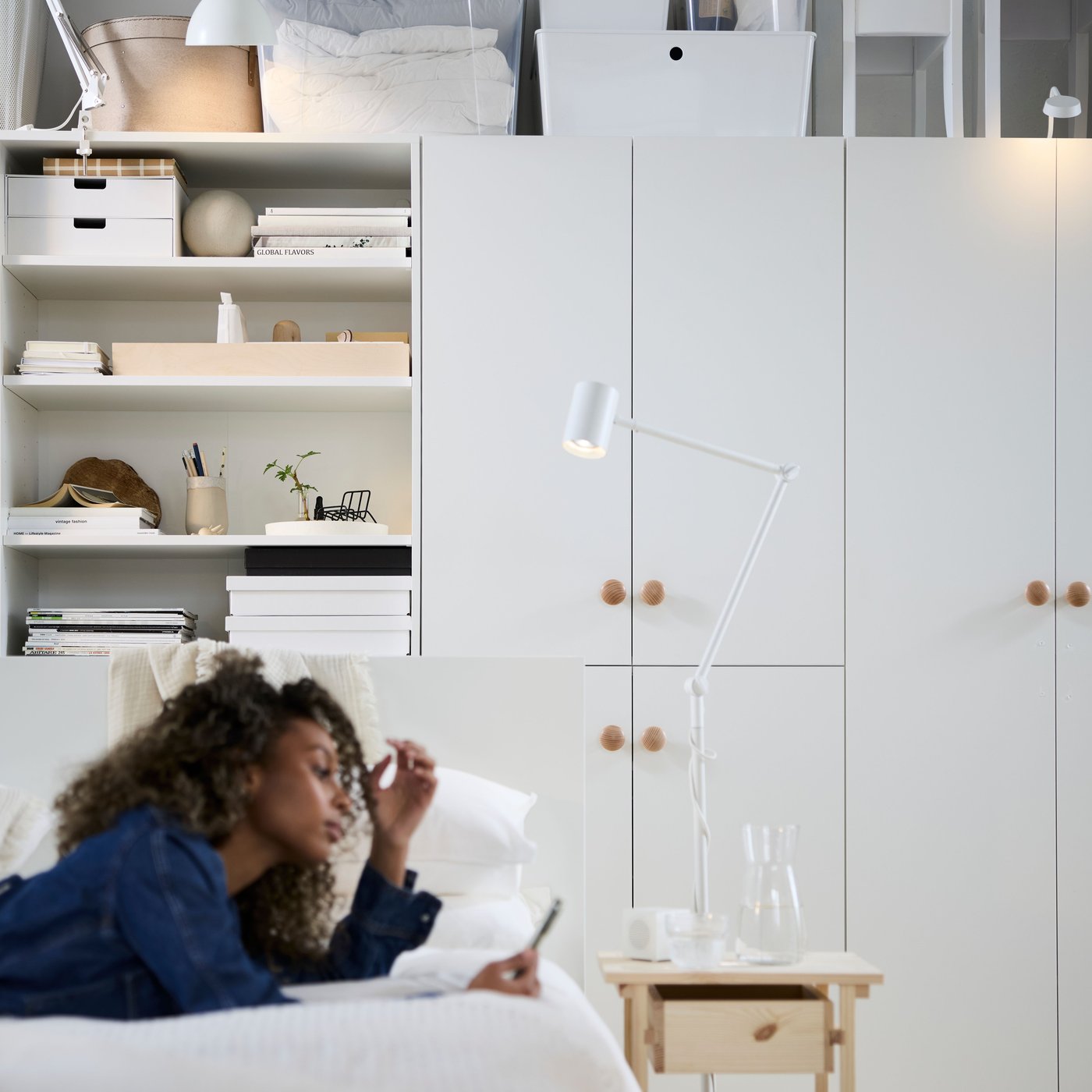 Une personne se détend sur un lit dans un espace de vie multifonctionnel meublé d’étagères et rangements LASTARE blancs.