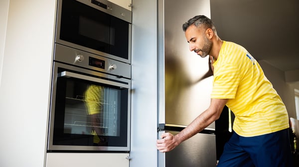 A person in a yellow striped work shirt installs a built-in kitchen appliance, adjusting a tall cabinet next to a stainless steel oven in a modern kitchen.