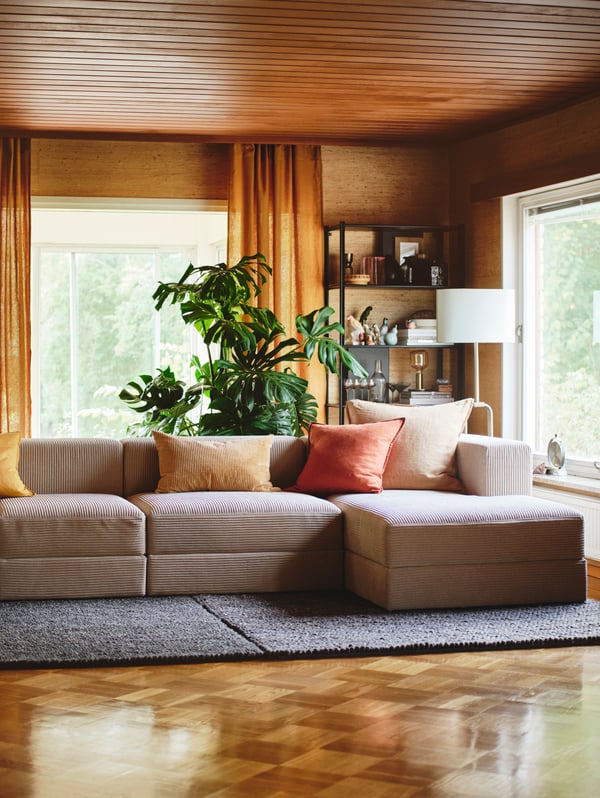 A living room with a grey/beige JÄTTEBO 4.5-seat modular sofa with chaise longue with cushions on it, and a big plant behind.