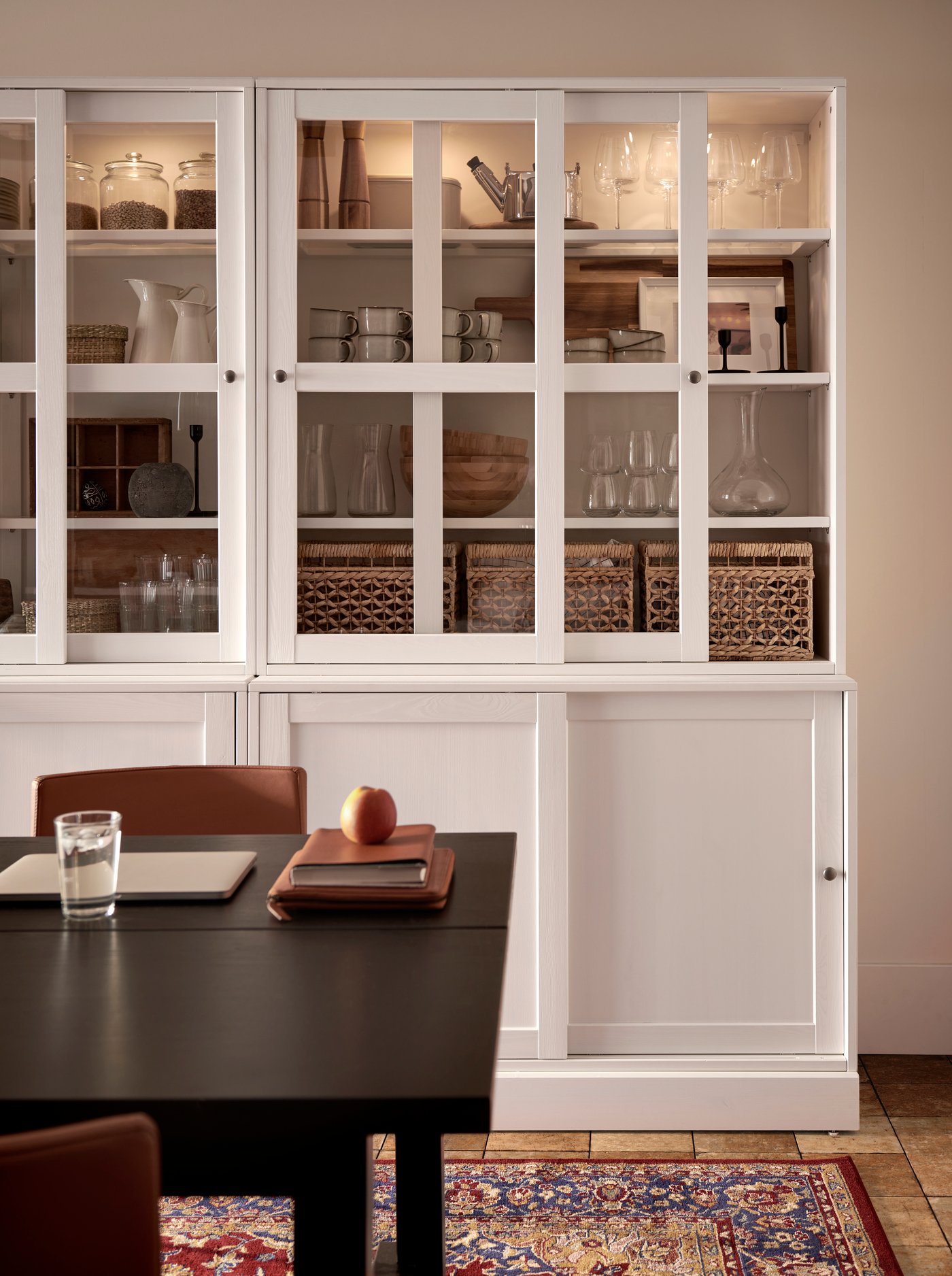 Une salle à manger avec une table noire sur laquelle reposent un verre d’eau, un ordinateur portable fermé et quelques livres, avec en arrière-plan une vitrine blanche remplie de verres, bols et paniers, sur un tapis à motifs.