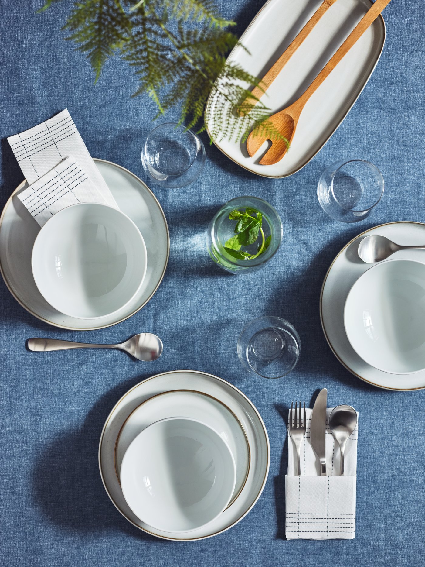 Vista superior de una mesa puesta con platos y bowls OFTAST blancos sobre un mantel azul claro, vasos transparentes y cubiertos plateados. 