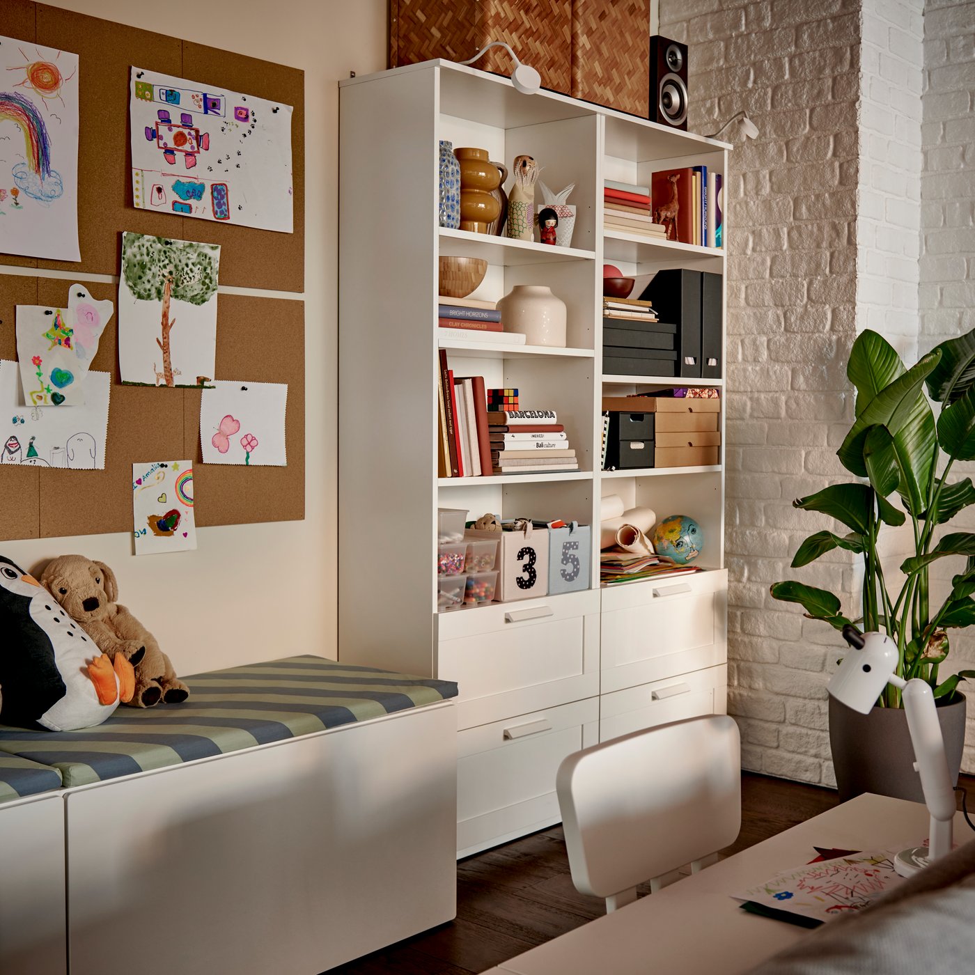 Una estantería blanca BRIMNES, repleta de libros y cajas decorativas, junto a un acogedor rincón de lectura con animales de peluche.