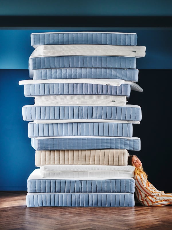 In a blue room, a person in striped pyjamas is sitting on the floor and leaning against a stack of IKEA mattresses.