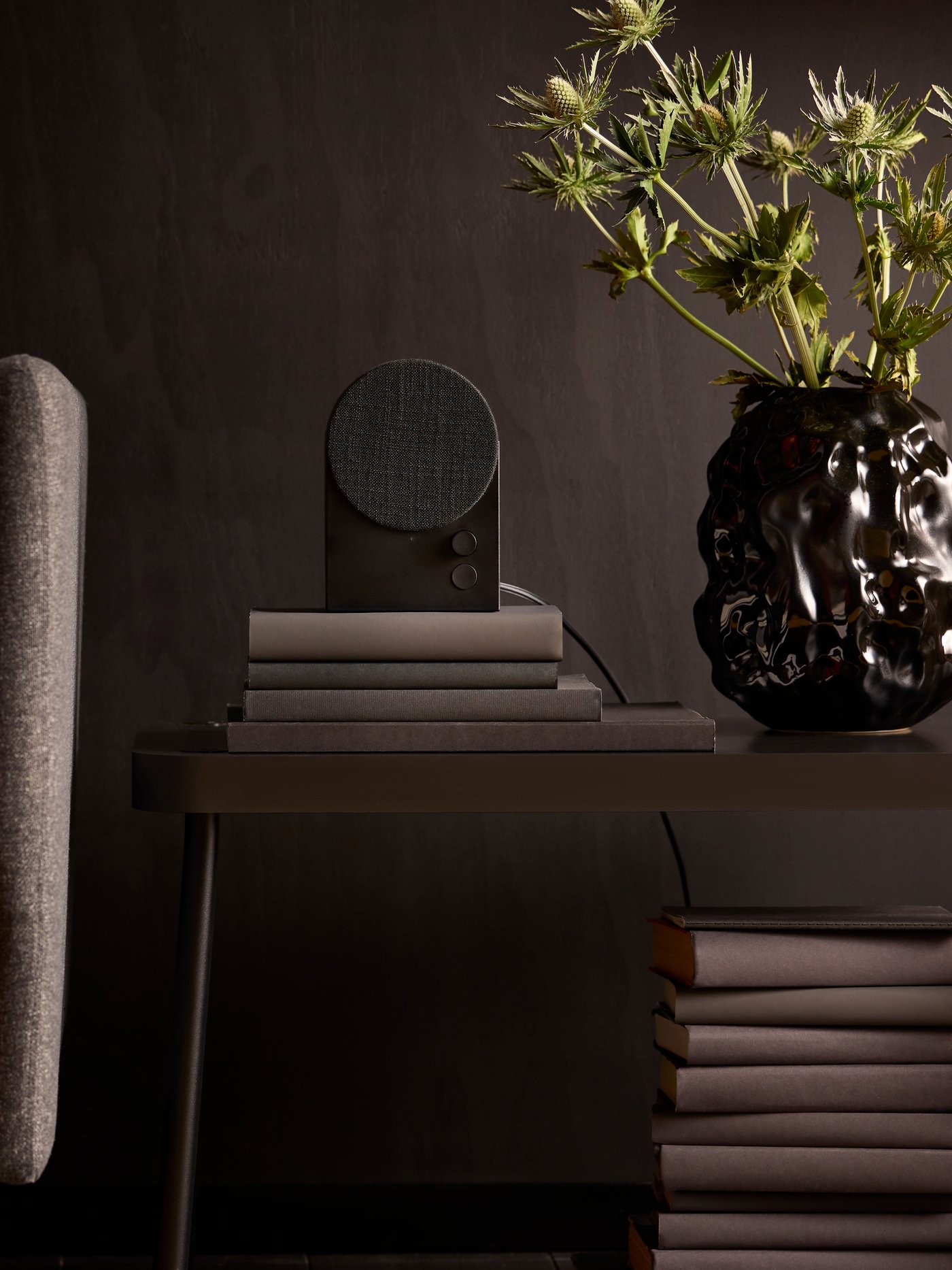 A black NATTBAD Bluetooth speaker sitting on a stack of books on a table beside a dark grey armchair.