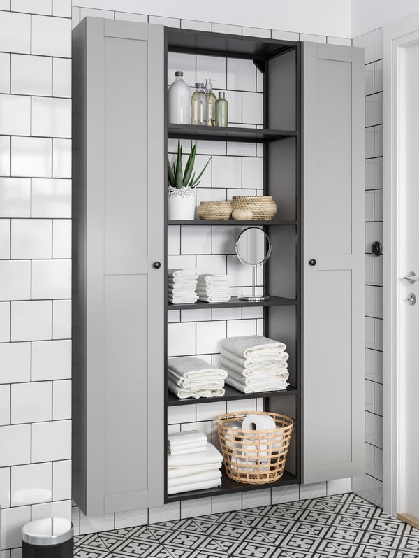 Bagno piastrellato di bianco con due mobili alti in grigio e, in mezzo, uno scaffale nero.