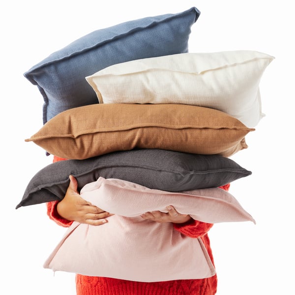 Someone holds a stack of cusions in their arms, covering their face. Each cushion has a different coloured LAGERPOPPEL cover.