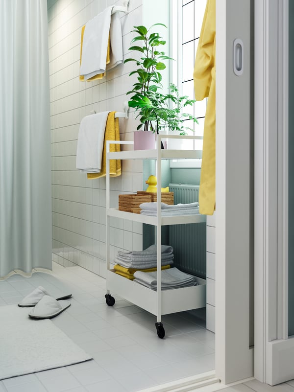 A white trolley with castors with two plants on the top shelf and wooden boxes and towels on the lower shelves.