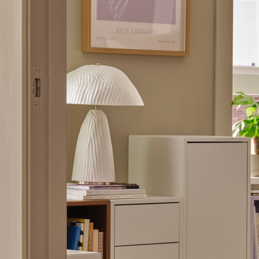 Vit VINDKRAFT bordslampa med veckad struktur står på ett skåp bredvid böcker och en tavla på väggen, i ett ljust och modernt rum.