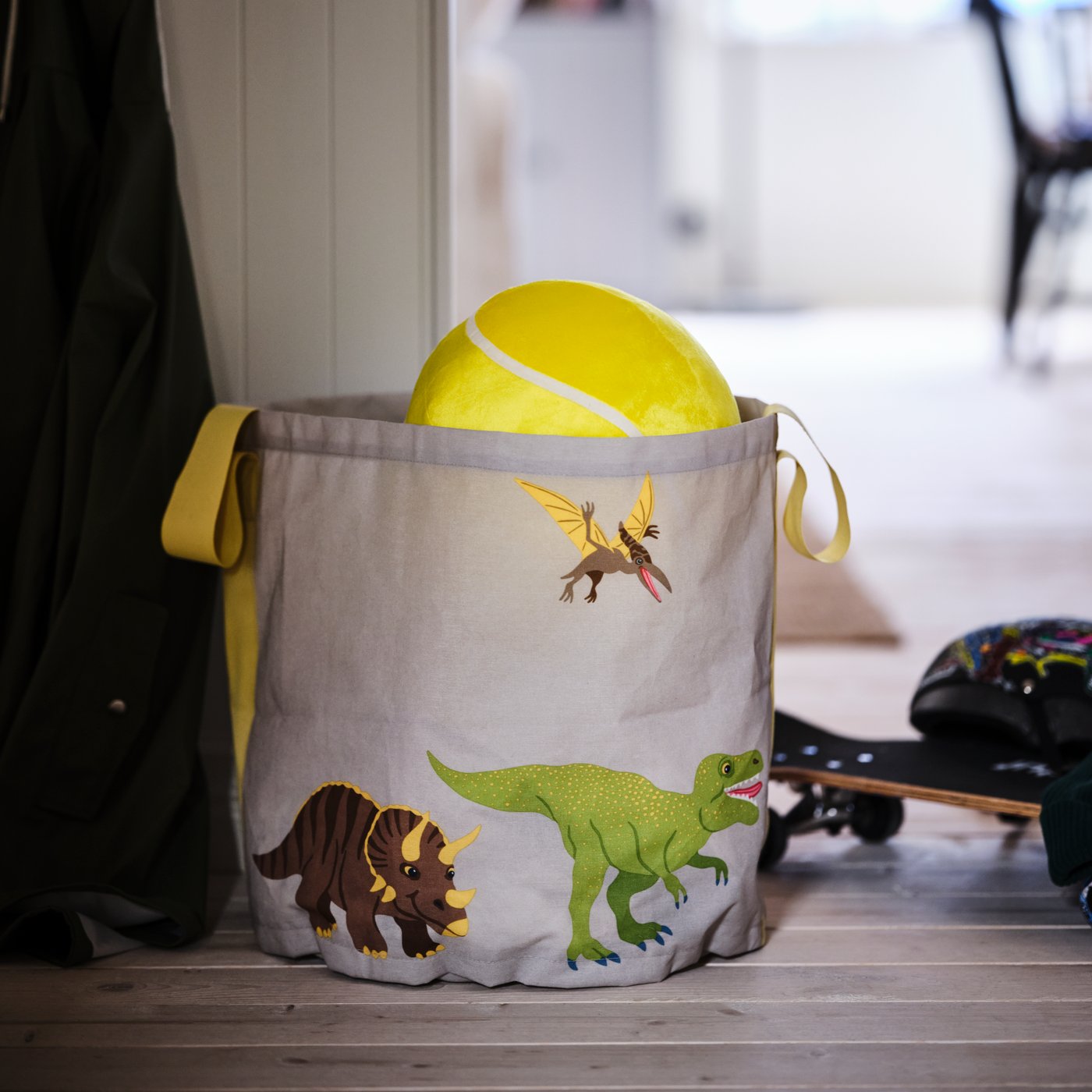Na dřevěné podlaze chodby stojí úložná taška JÄTTELIK se vzorem dinosaurů s hračkami uvnitř a kolem ní.