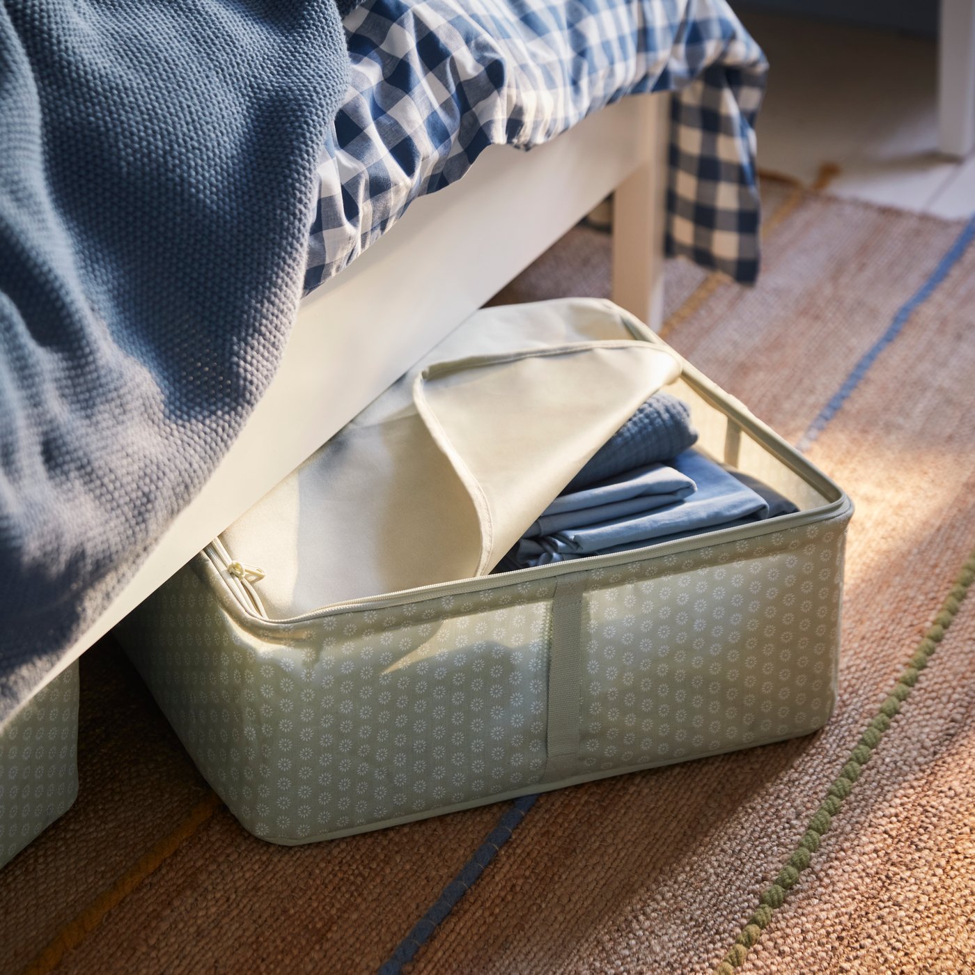 A pale grey-green HEMMAFIXARE storage box filled with blue bedding sticks out from under a white bed.