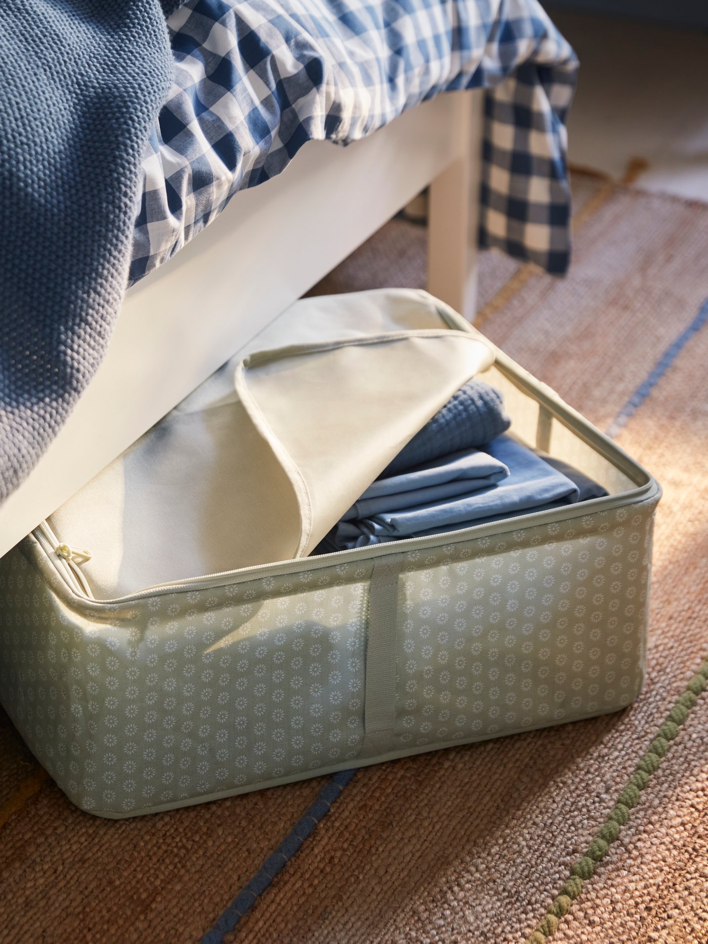 A pale grey-green HEMMAFIXARE storage box filled with blue bedding sticks out from under a white bed.