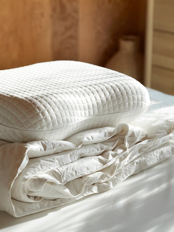 Plumón de plumas FJÄLLARNIKA doblado sobre una cama, con una almohada KLUBBSPORRE encima.